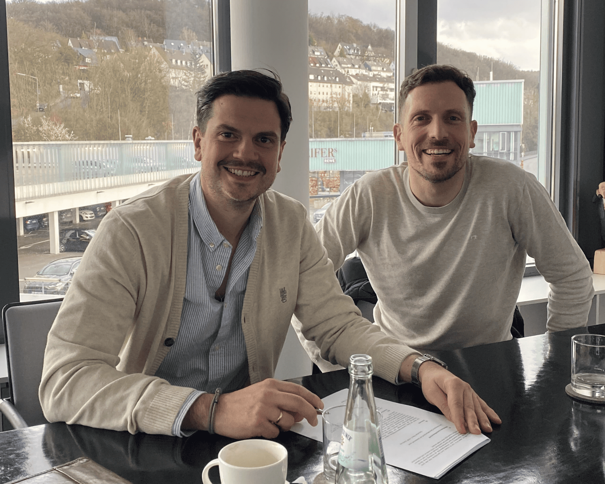 Zwei Personen sitzen an einem Tisch mit Dokumenten und lächeln in die Kamera
