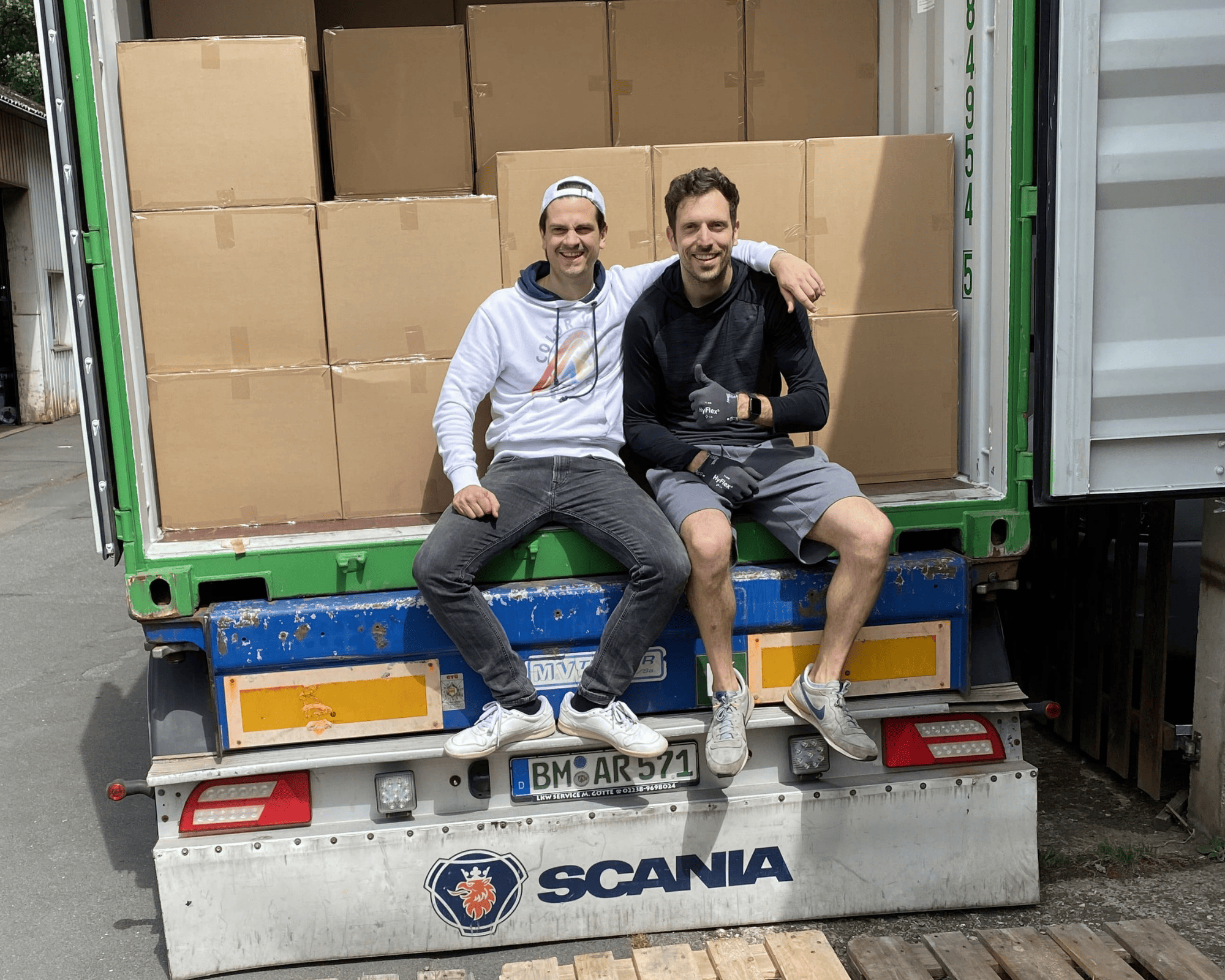Zwei Personen sitzen auf der Ladeklappe eines LKWs voller Kartons und lachen in die Kamera